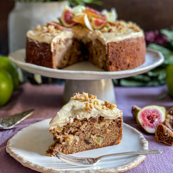 A rustic Fig and Walnut Cake sits on a wooden cutting board, adorned with a glossy honey glaze drizzled over the top. Chopped walnuts and thin slices of fresh figs decorate the surface, adding texture and charm. The soft, dense crumb is visible through a slice, showcasing its delicate texture and vibrant flavors.